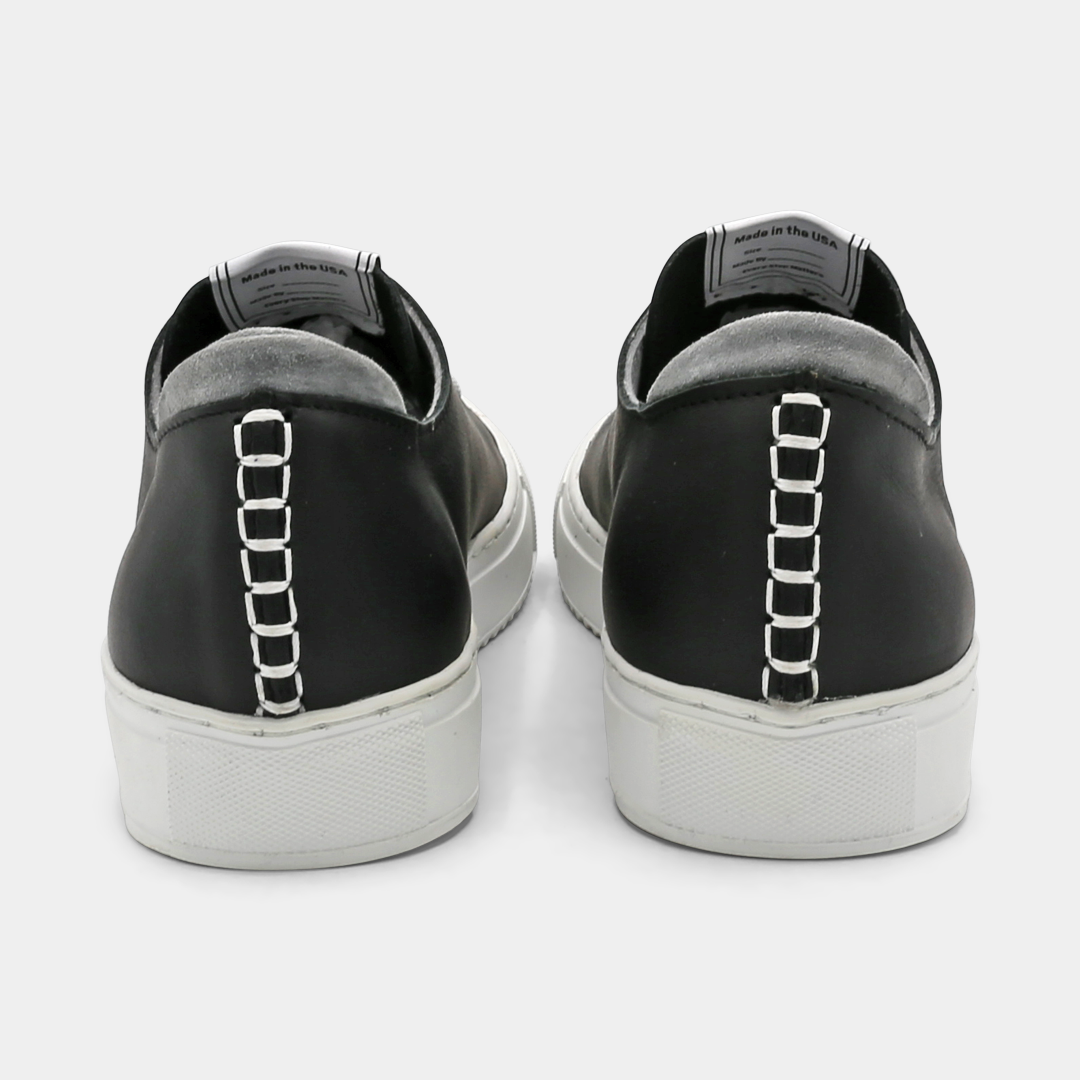 A black colored low-top sneaker with a white sole and laces, back view.