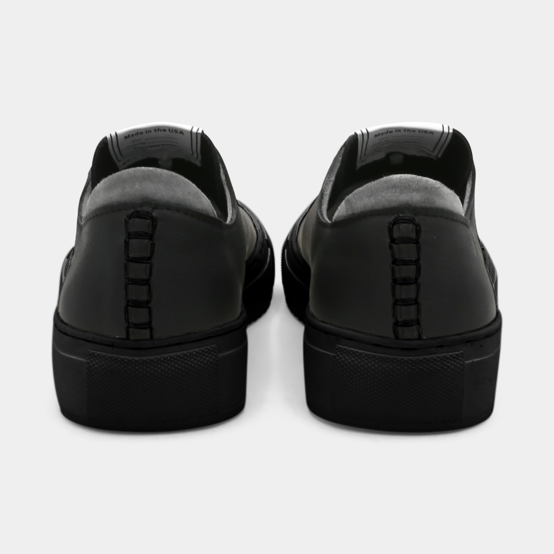A black colored low-top sneaker with a black sole and laces, back view.