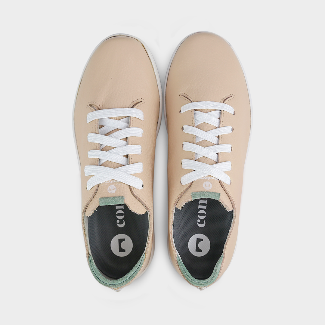 A top view shot of pair of nude beige leather sneakers with a lace-up closure featuring flat white shoelaces, white printing on the tongue and a dark grey sockliner with a circle logo and COMUNITY printed in white with a green heel accent.