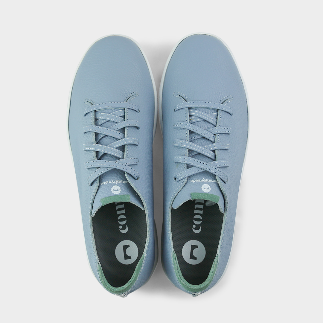 A top view shot of pair of sky blue leather sneakers with a lace-up closure featuring flat sky blue leather shoelaces, white printing on the tongue and a dark grey sockliner with a circle logo and COMUNITY printed in white with a green heel accent.