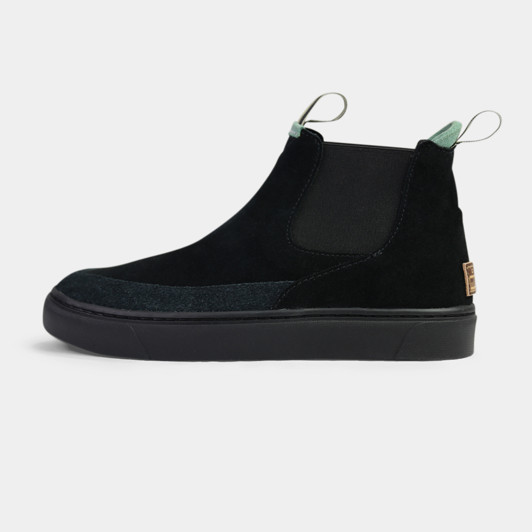 A side-shot of a pair of black suede boots. The shoes have black elastic gussets on the side and a striped pull tab at the heel. The black insoles feature a white