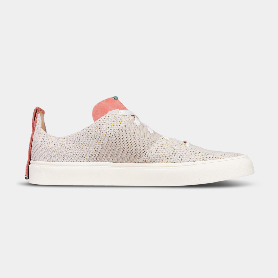 A pair of off-white knitted sneakers with light pink speckles. The shoes have white laces, a pale pink tongue, and a pink and white heel loop. The insoles are dark green with a white text logo and a circle.