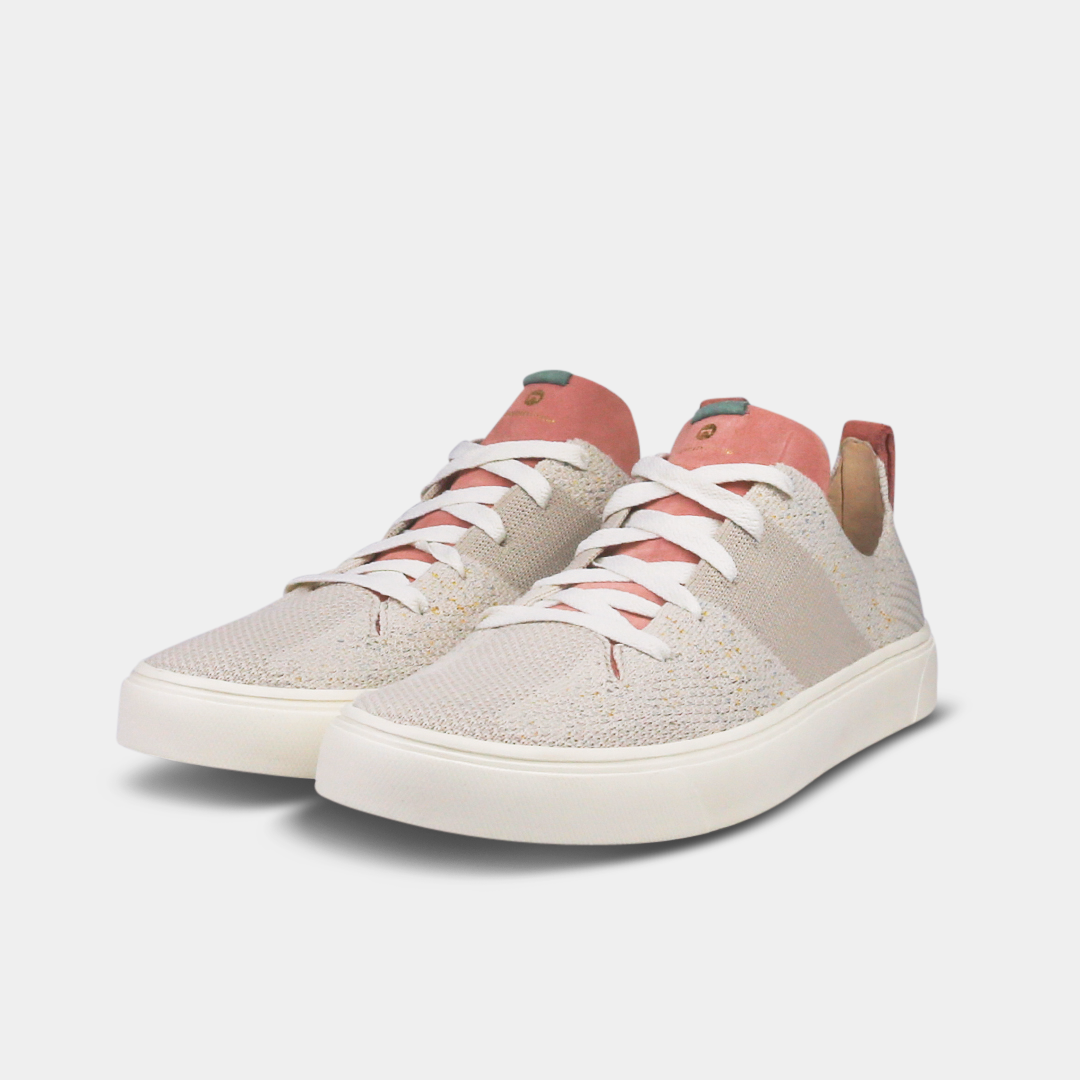 A pair of off-white knitted sneakers with light pink speckles. The shoes have white laces, a pale pink tongue, and a pink and white heel loop. The insoles are dark green with a white text logo and a circle.