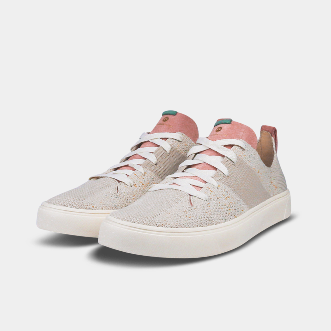 A pair of off-white knitted sneakers with light pink speckles. The shoes have white laces, a pale pink tongue, and a pink and white heel loop. The insoles are dark green with a white text logo and a circle.