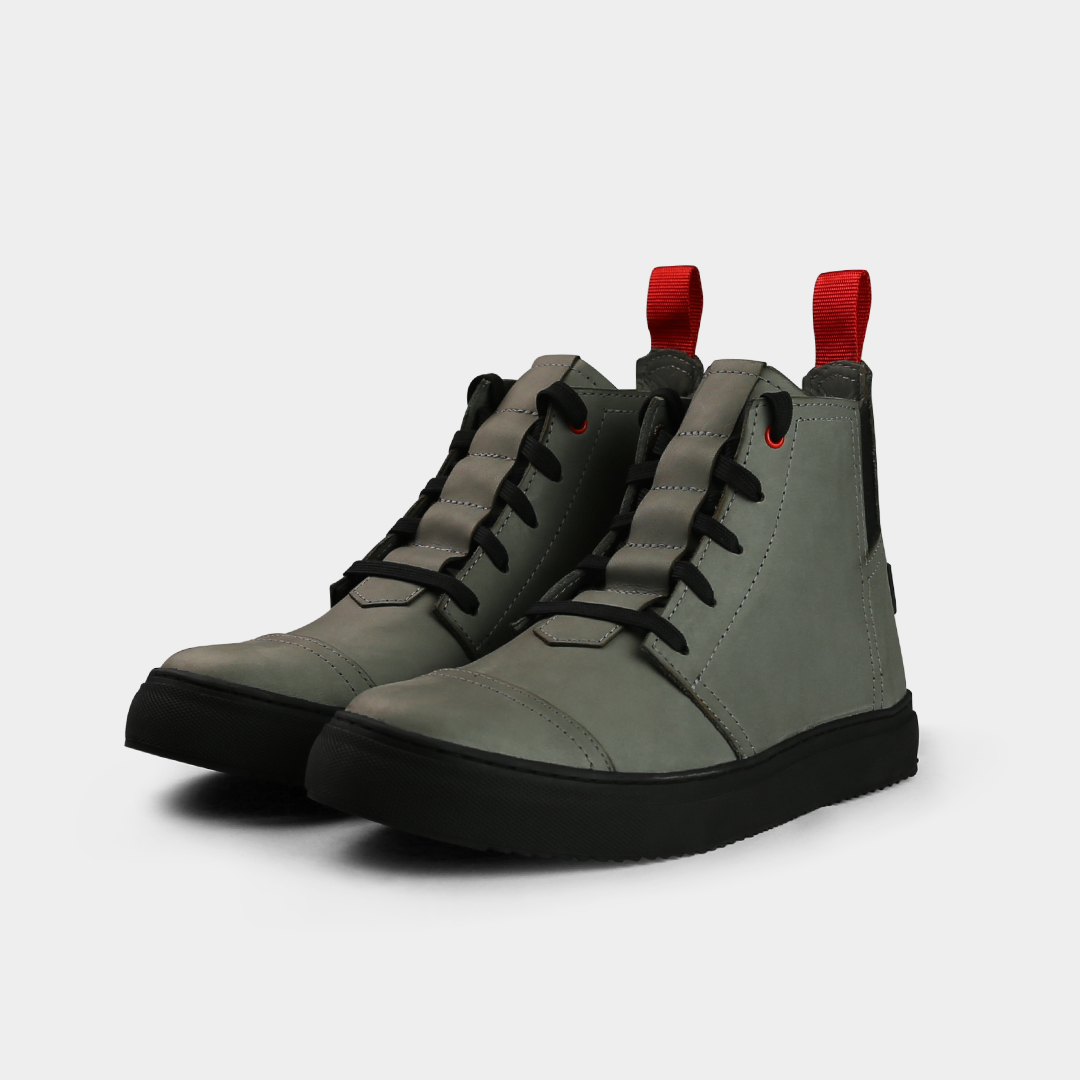 A low-angle product shot of a pair of grey high-top shoes, placed side-by-side. The shoes are made of grey leather, with black laces and a thick black sole. A bright red loop is attached to the back of each heel. The shoes have a sleek, modern design, and they are on a solid light gray background.
