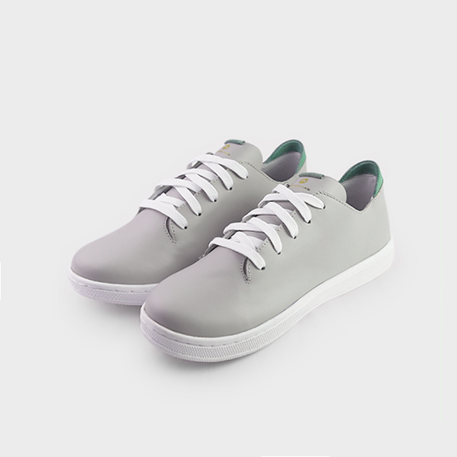 A quarter angle shot of pair of light grey leather sneakers with a lace-up closure featuring flat white shoelaces, a white rubber cupsole and a green accent on the heel.