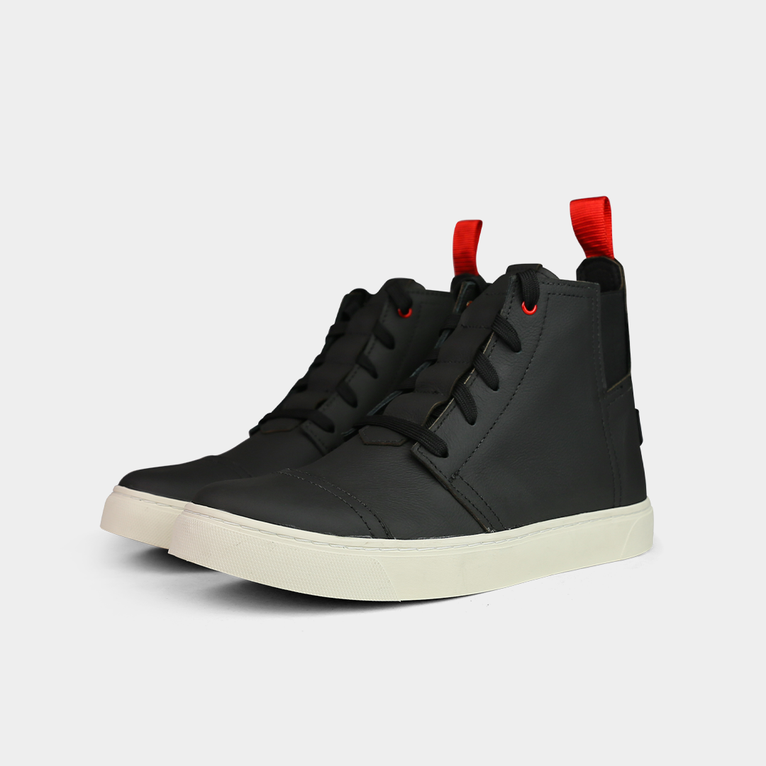 A low-angle product shot of a pair of black high-top shoes, placed side-by-side. The shoes are made of black leather, with black laces and a thick black sole. A bright red loop is attached to the back of each heel. The shoes have a sleek, modern design, and they are on a solid light gray background.