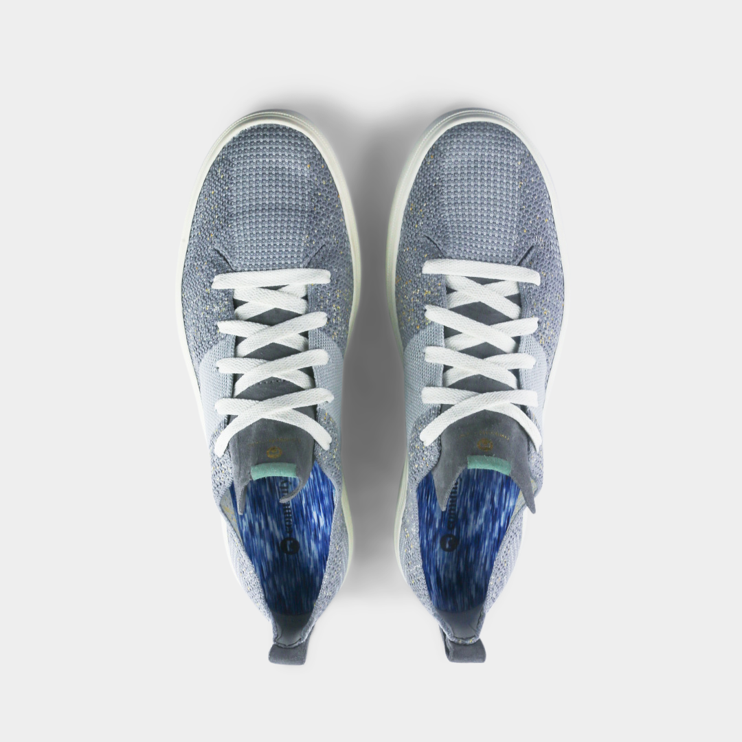 A pair of light gray knitted sneakers. The shoes have a thick, gray suede pull tab on the back with a gold logo and a small mint green detail at the base.