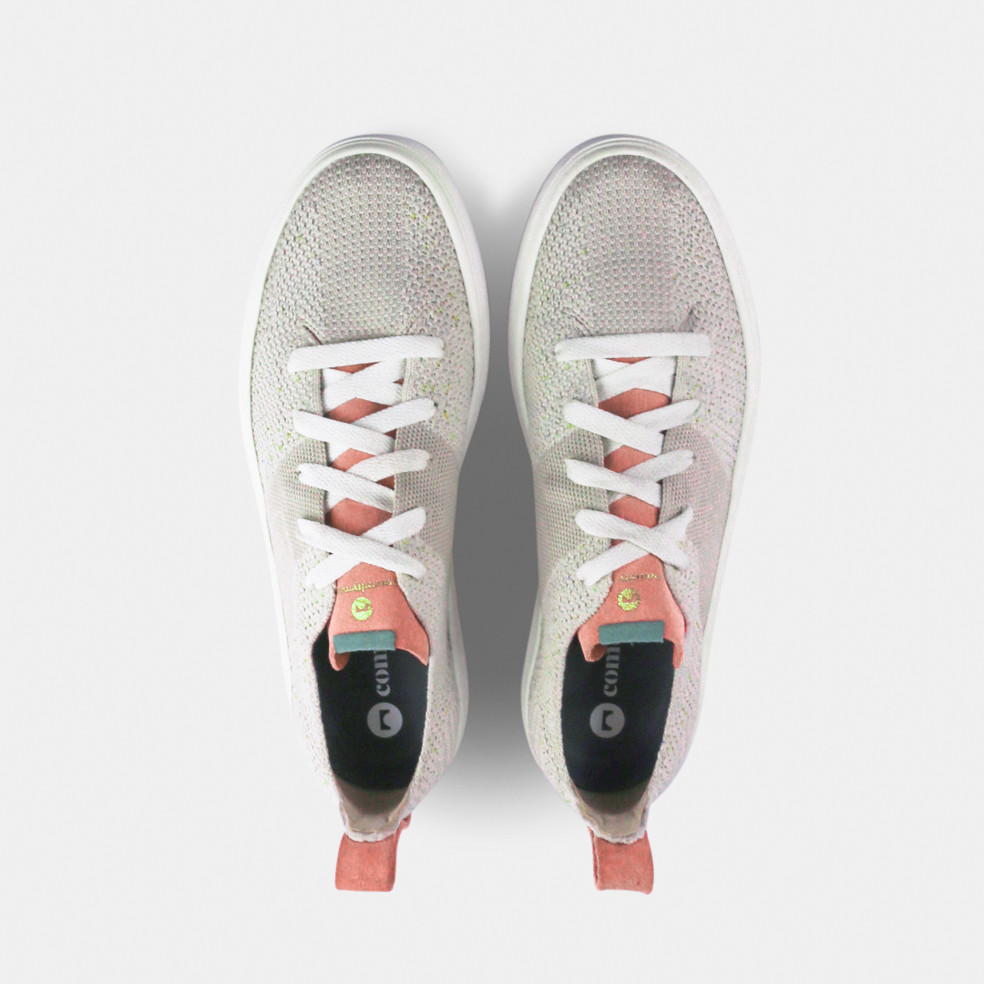 A pair of off-white knitted sneakers with light pink speckles. The shoes have white laces, a pale pink tongue, and a pink and white heel loop. The insoles are dark green with a white text logo and a circle.