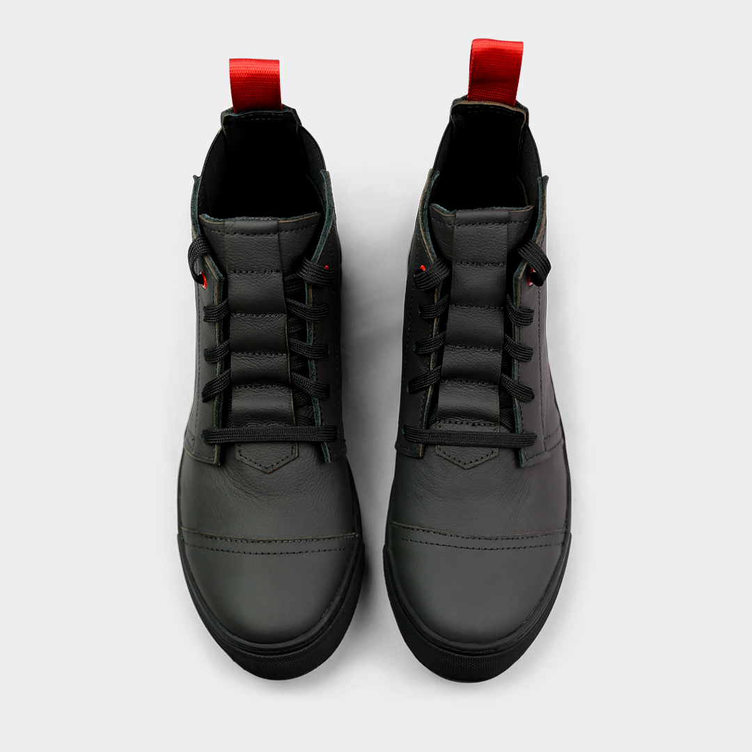 A high-angle, top-down product shot of a pair of black high-top shoes. The shoes are made of black leather with black laces, a small red accent on the eyelets, and a thick black sole. A red loop is visible on the back of each heel. The shoes are on a solid light gray background.
