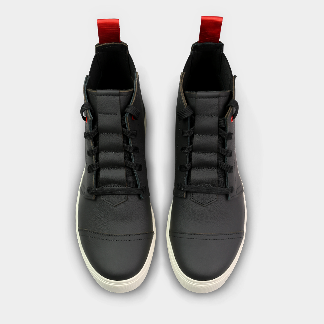 A high-angle, top-down product shot of a pair of black high-top shoes. The shoes are made of black leather with black laces, a small red accent on the eyelets, and a thick black sole. A red loop is visible on the back of each heel. The shoes are on a solid light gray background.