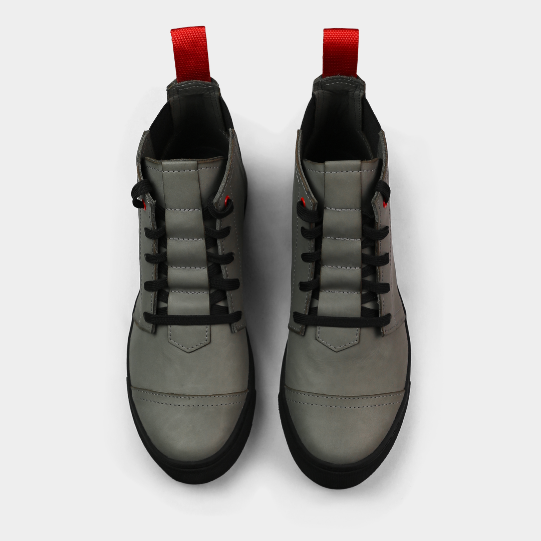 A high-angle, top-down product shot of a pair of grey high-top shoes. The shoes are made of black leather with black laces, a small red accent on the eyelets, and a thick black sole. A red loop is visible on the back of each heel. The shoes are on a solid light gray background.