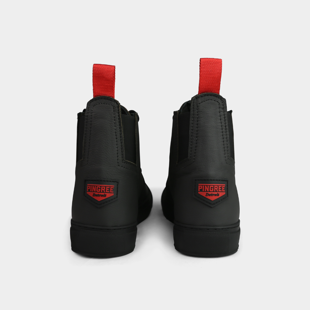 A head-on, product shot of the back of a pair of black high-top shoes. The shoes are made of black leather, with a white sole and a red loop on the back of the ankle. A red square with