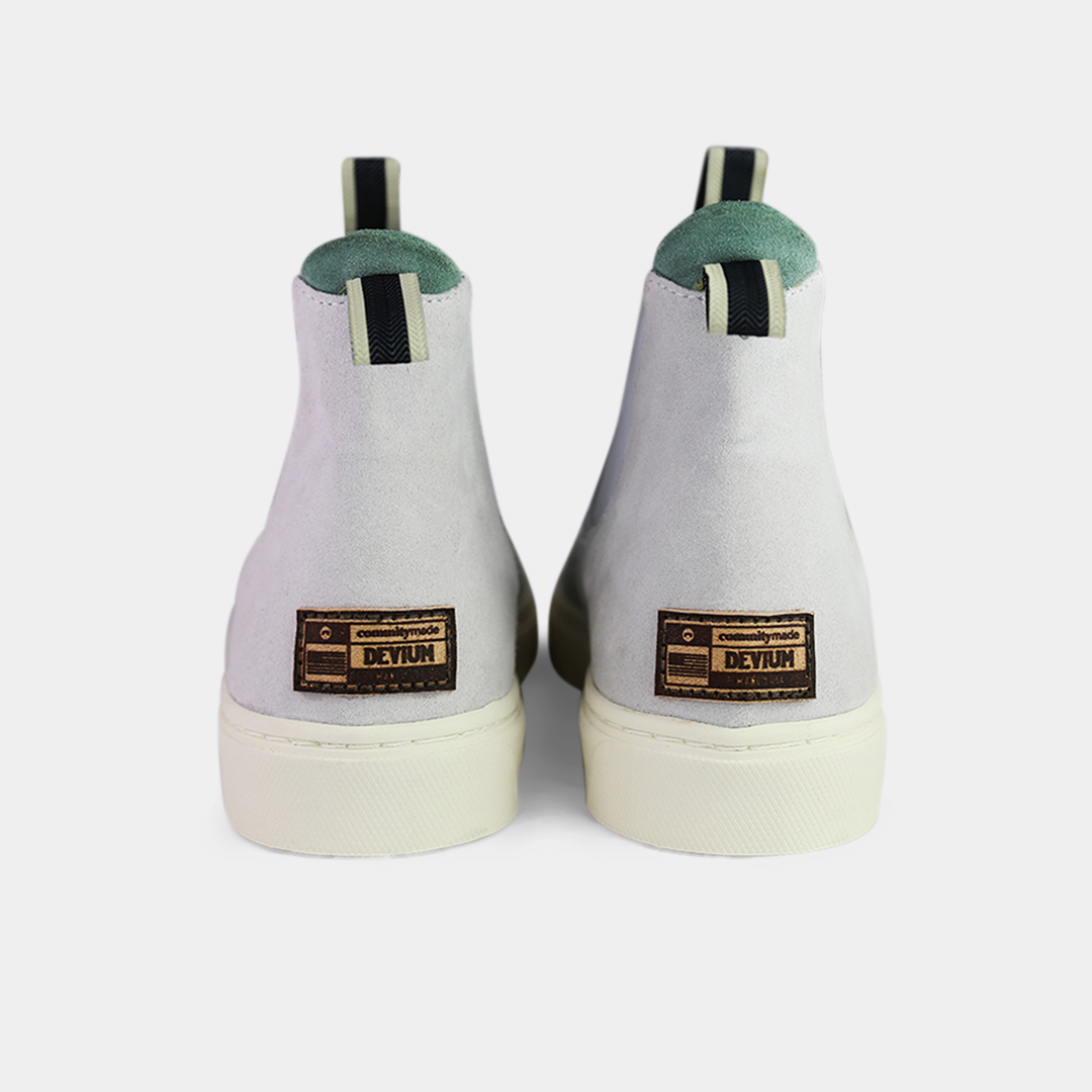 A pair of off-white suede ankle boots with an off-white sole. The boots feature a light brown leather logo tag on the back of each heel, a mint green elasticated panel, and a black and white striped pull tab at the back of the ankle.