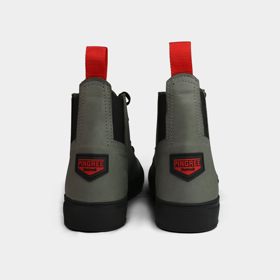 A head-on product shot of the back of a pair of gray high-top shoes. The shoes are made of gray leather, with a thick black sole and a red loop on the back of the ankle. A red and black shield-shaped patch with