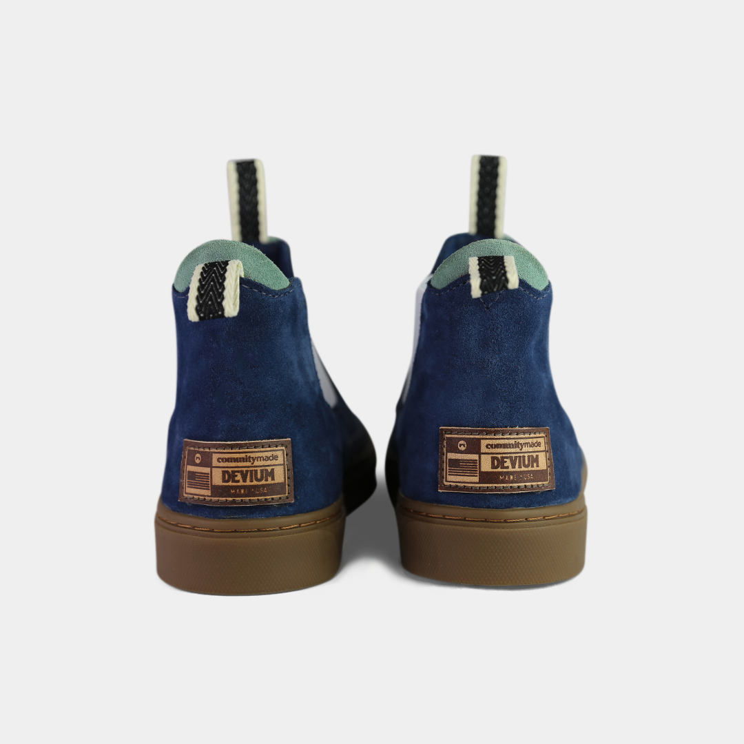 A pair of dark blue suede ankle boots with a brown rubber sole. The boots feature a light brown leather logo tag with