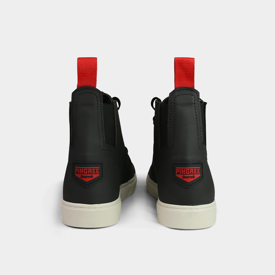 A head-on, product shot of the back of a pair of black high-top shoes. The shoes are made of black leather, with a white sole and a red loop on the back of the ankle. A red square with