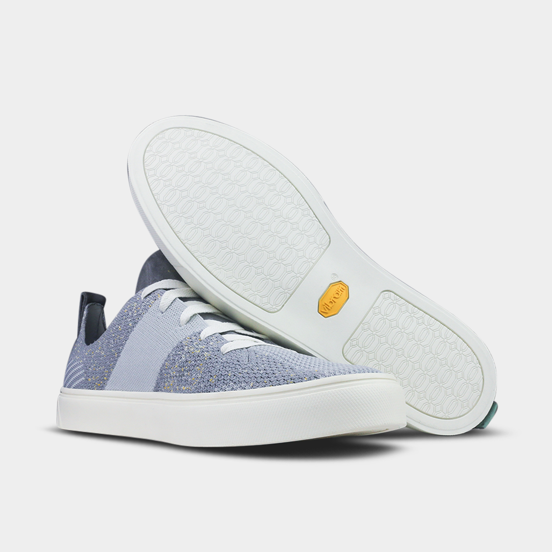 A knitted light gray and white sneaker with a chunky white sole and white laces. The shoe has a thick white stripe down the side. Another shoe is shown, turned upside down to show the white rubber sole with a honeycomb pattern and a yellow logo.