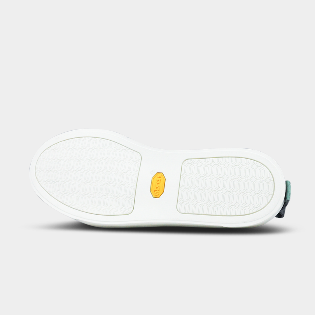 The bottom of a shoe with a white rubber sole. The front and back of the sole have a repeating, circular pattern for grip, with a bright yellow oval logo in the center. A small dark green piece of material is visible at the heel