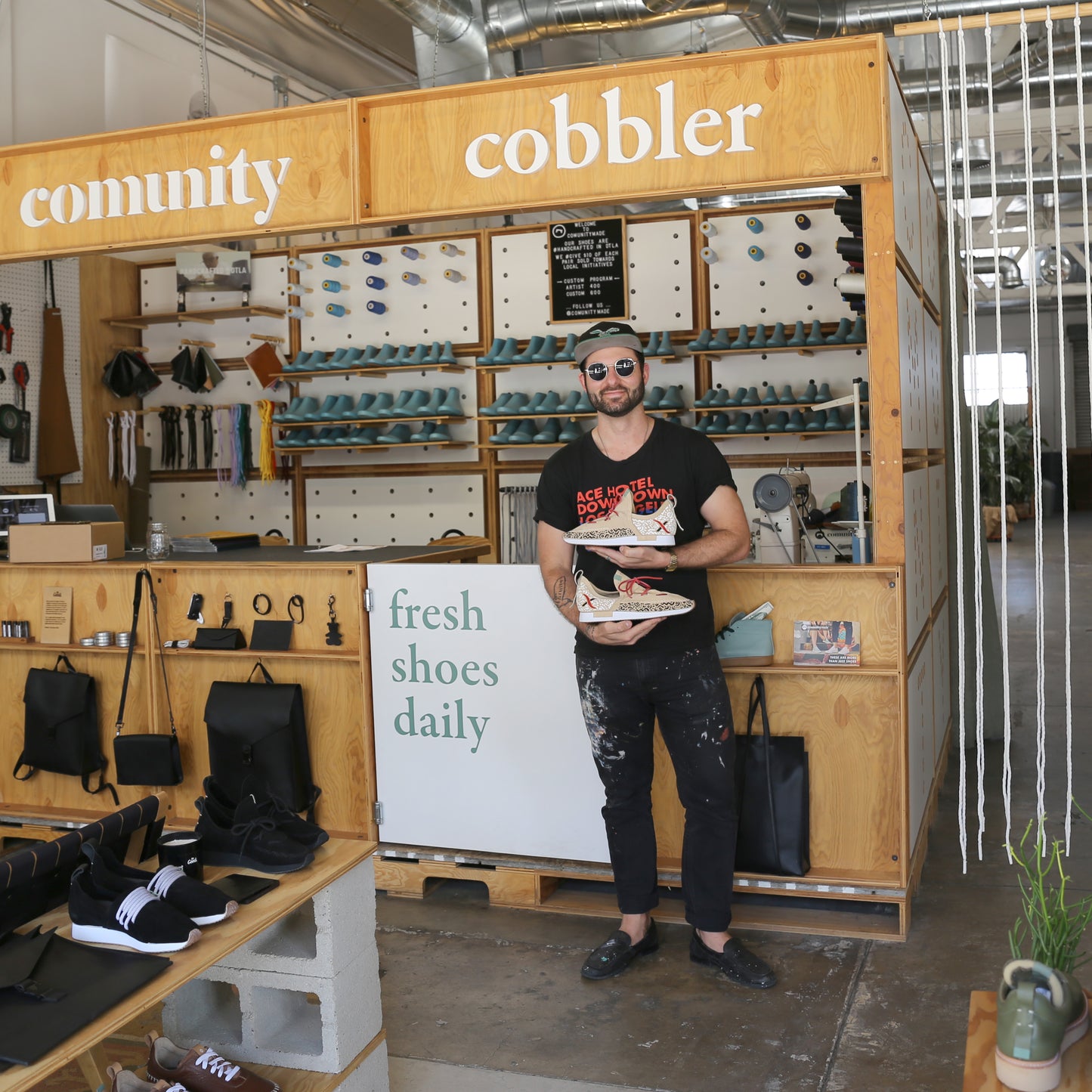 Photo of a male artist standing in front of a wood structure that says COMUNITY Cobbler holding a pair of beige suede custom art shoes that he has painted. He is wearing black pants, black shirt and  black hat and is standing in a retail showroom with shoes and bags.