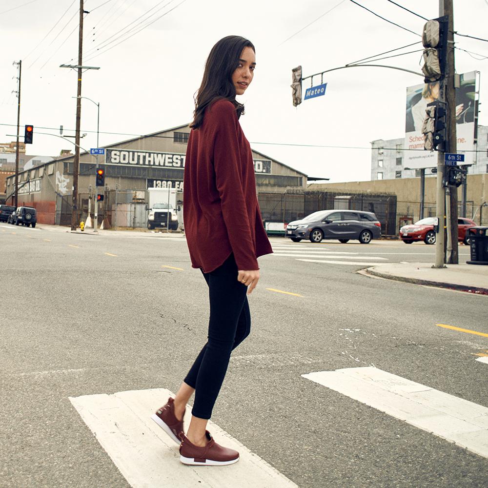 A woman with dark hair, wearing a loose-fitting burgundy sweater, black pants, and Comunitymade burgundy leather shoes with white soles. She is walking across a crosswalk in an urban setting with a large industrial building, streetlights, and cars in the background.