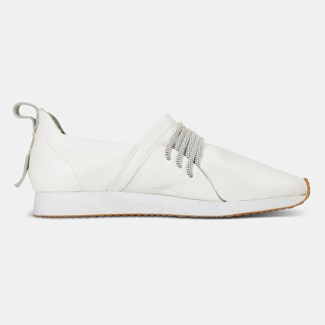 A side profile of a single white Comunitymade slip-on shoe. The shoe is made of a white, pebbled leather with thick gray and white speckled laces at the top. It has a flat white sole with a gum-colored accent at the toe.