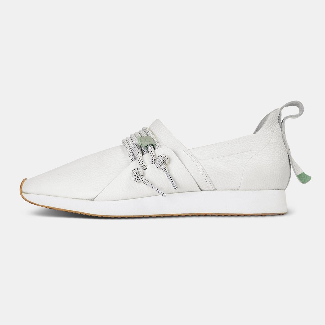 A side profile of a single white Comunitymade slip-on shoe. The shoe is made of a white, pebbled leather with thick gray and white speckled laces at the top. It has a flat white sole with a gum-colored accent at the toe.