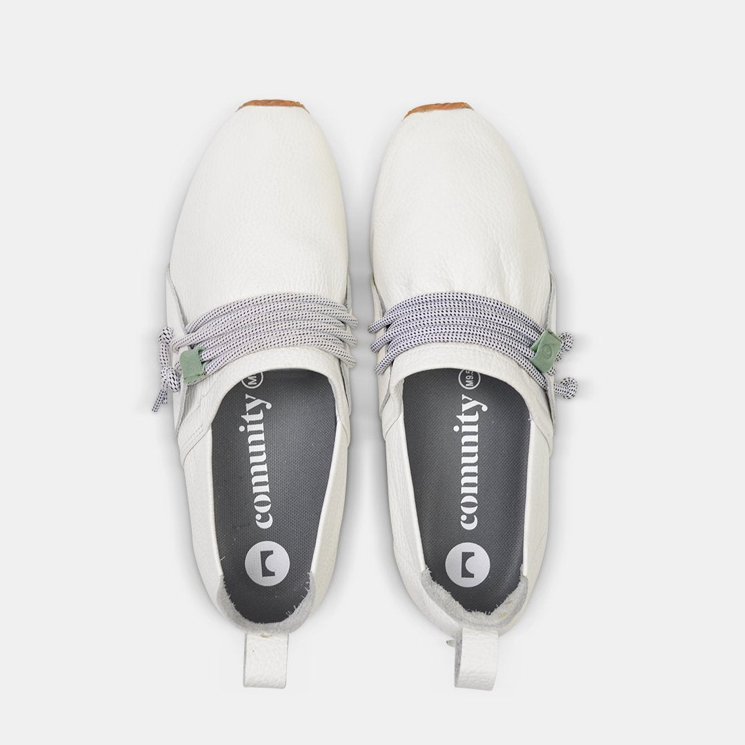 A top-down view of two white Comunitymade slip-on shoes. The shoes are made of a white, pebbled leather with thick gray and white speckled laces that wrap around the top. The insoles are dark gray with