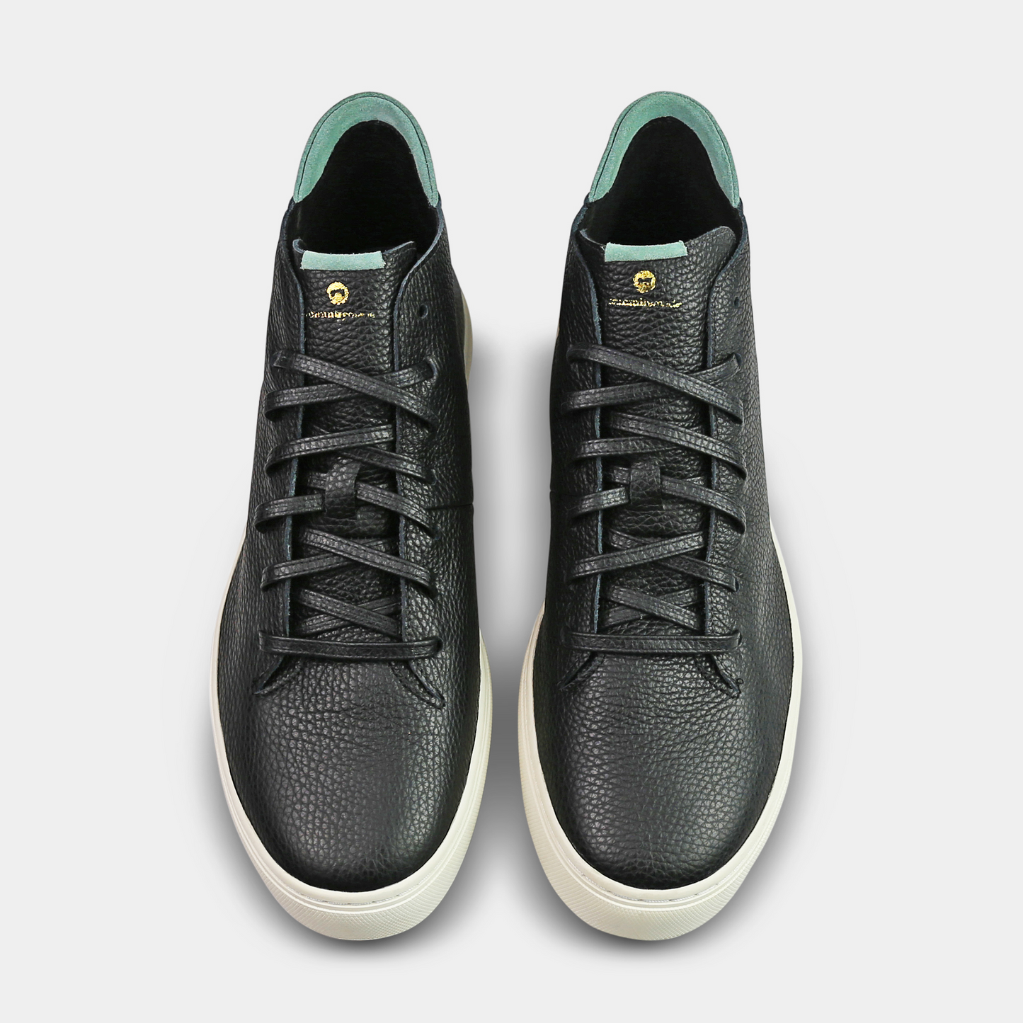 A high-angle product shot of a pair of black high-top sneakers, viewed from directly above. The shoes are made of what appears to be pebbled leather and have black laces and a light-colored sole. The inside lining and the back of the tongue are a light green or mint color. The shoes have a small gold accent near the top of the laces, and one shoe has a small blue ring on one of the eyelets. The background is a solid light gray.