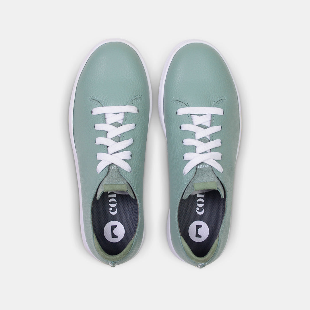 A top view shot of pair of light green leather sneakers with a lace-up closure featuring flat white shoelaces, white printing on the tongue and a dark grey sockliner with a circle logo and COMUNITY printed in white with a green heel accent.