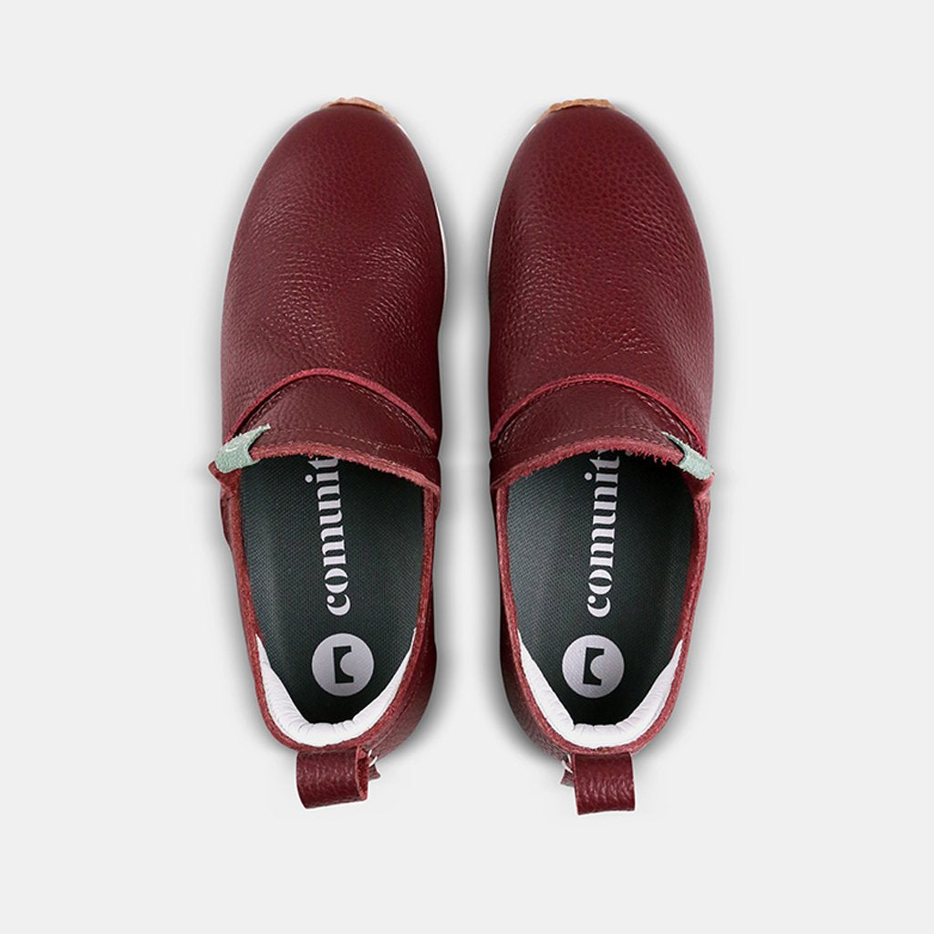 A top-down view of two Comunitymade slip-on shoes. The shoes are made of a deep burgundy, pebbled leather with a mint green accent loop at the heel. The insoles are dark gray with