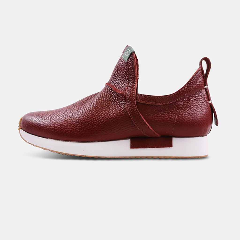 A side profile of a single Comunitymade sneaker-boot with a high-cut design. The shoe is made of a deep burgundy, pebbled leather. It has a thick white sole, a pull tab at the back, and the text