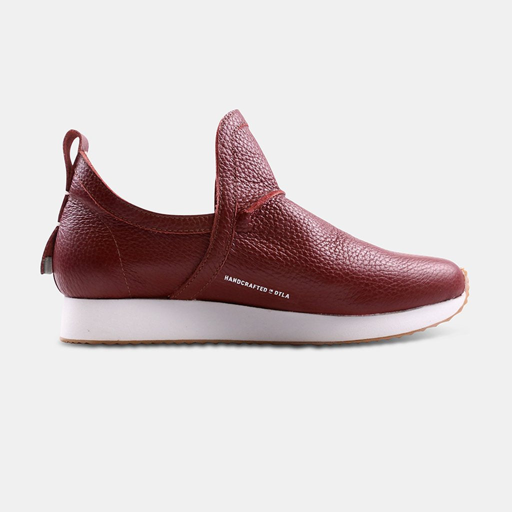 A side profile of a single Comunitymade sneaker-boot with a high-cut design. The shoe is made of a deep burgundy, pebbled leather. It has a thick white sole, a pull tab at the back, and the text