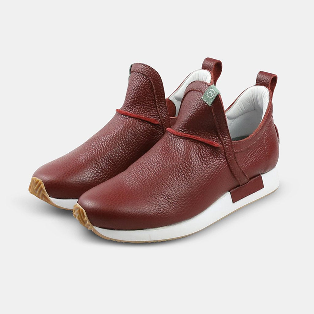 A front, three-quarter view of two Comunitymade sneaker-boots. The shoes are a deep burgundy, pebbled leather with a high-cut design and white soles. They feature a red cord detail on the tongue and a small mint green accent piece near the laces.