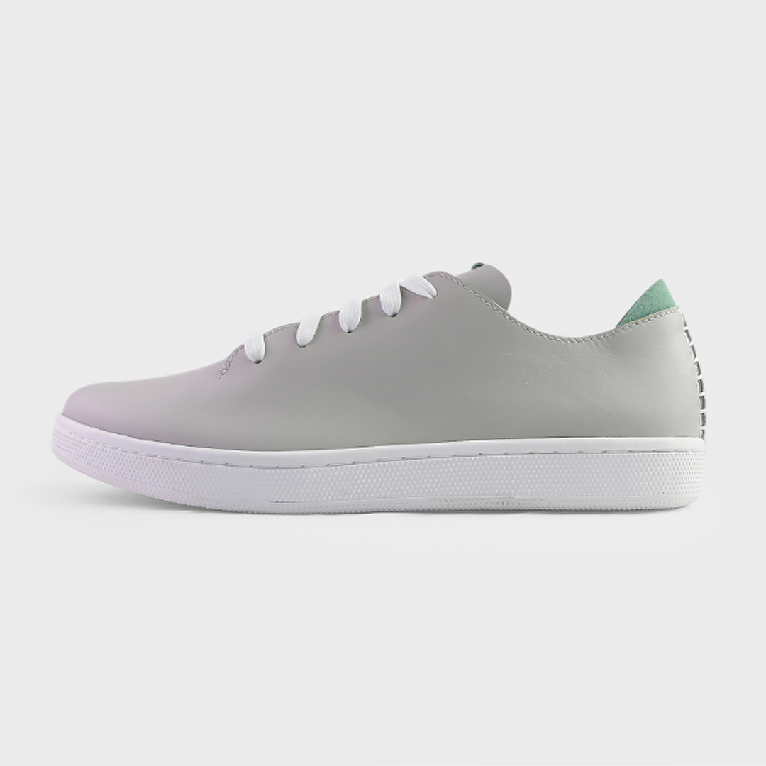 A profile shot of single light grey leather sneaker with a lace-up closure, featuring flat white shoelaces, a white rubber cupsole and a green accent on the heel.