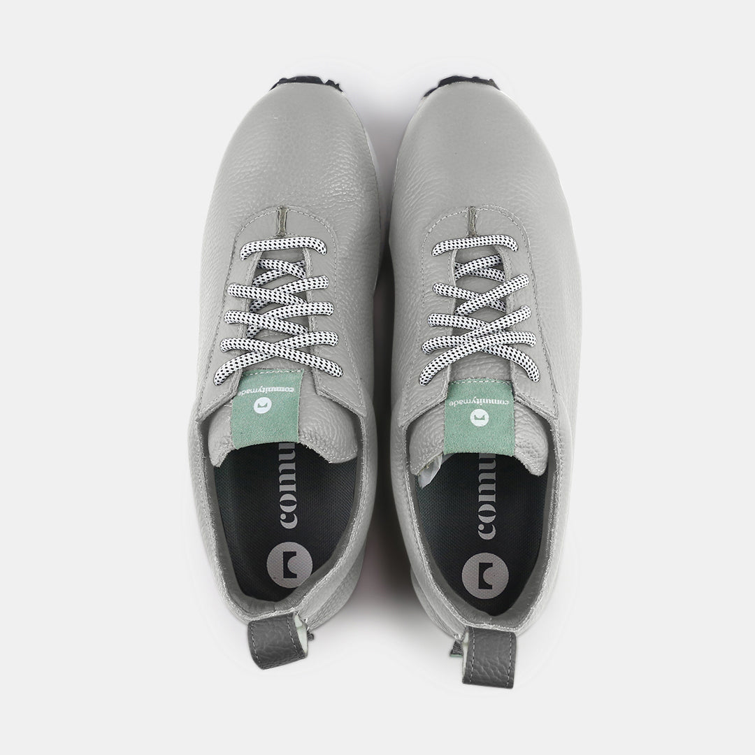 A pair of gray sneakers with a subtle grainy texture and white soles. A small mint green tag is at the base of the heel, just above the sole. A small white text logo is printed vertically on the back of each shoe.