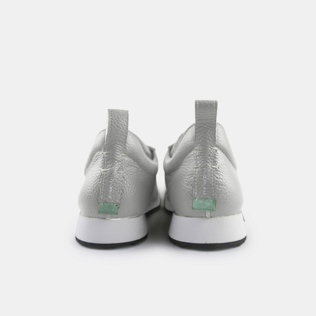 A pair of gray sneakers with a subtle grainy texture and white soles. A small mint green tag is at the base of the heel, just above the sole. A small white text logo is printed vertically on the back of each shoe.
