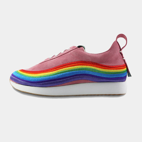 The shoe has a thick, white platform sole and is decorated with a vibrant, embroidered rainbow stripe that curves along the side of the shoe, from the toe to the heel. It has white laces and a pink loop on the back of the heel. The sneaker is positioned on a solid gray background.