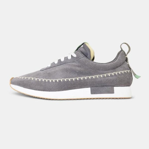A side profile of a single grey Comunitymade lace-up shoe. The shoe is made of a grey suede, has flat white laces and white accent stitching around the entire shoe, plus a grey heel pull tab. It has a flat white midsole with a gum-rubber colored outsole.